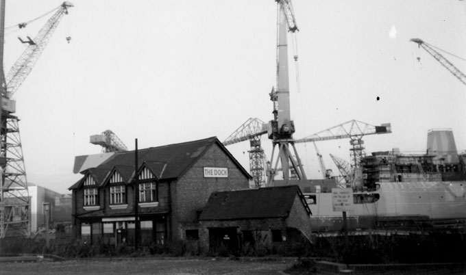 B&W photo of the Dock pub in Wallsend, Tyne and Wear, taken in the late 1980s. A ship and several of the iconic Swan Hunter shipyard cranes tower above the pub.
Andrew Green